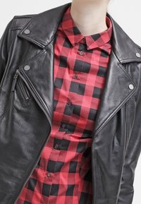Giubbotto da moto in pelle nera con hardware argentato, indossato sopra una camicia a scacchi rossi e neri con design a bottoni.