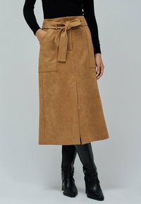 Suede midi sukně v odstínu tan s páskem v pase, bočními kapsami a předním rozparkem. Spárovaná s přiléhavými černými vysokými botami.