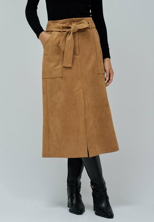 Rok midi suede berwarna coklat dengan pinggang berikat, saku samping, dan belahan depan. Dipasangkan dengan sepatu bot hak tinggi hitam yang pas.