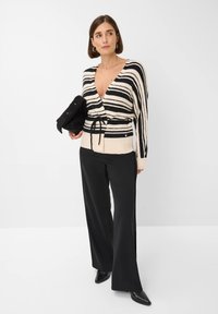 Gestreifter Strickcardigan in Creme und Schwarz mit tiefem V-Ausschnitt, akzentuiert mit einer Schleife an der Taille, kombiniert mit lockeren schwarzen Hosen und schwarzen Absätzen.