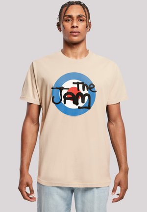 Beige Baumwoll-T-Shirt mit einem kreisförmigen blauen, weißen und roten Grafikdesign, auf dem "The Jam" in stilisiertem schwarzem Text steht. Lockerer Schnitt und kurze Ärmel.