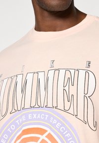 Lyserød bomulds t-shirt med stort sort blokbogstavstekst "SUMMER" og cirkulært grafisk motiv i orange og lilla detaljer. Korte ærmer, rund halsudskæring.