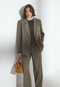 Ženska v olivno zeleni dvojno zapeti obleki, s pomočnim rjavim zgornjim delom in belim puloverjem s turtleneckom, drži majhno rjavo sueden torbico, stoji ob beli steni.