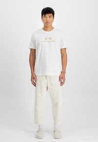 Bílý bavlněný tričko s zlatým logem "ALPHA INDUSTRIES", spárované s kalhotami volného střihu světlé barvy a šedými nazouvacími botami.