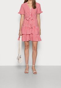 Robe vichy rouge et blanche avec des manches courtes, un nœud à l'avant et une jupe à volants. Associée à un sac bandoulière crème et des sandales à lanières beiges.