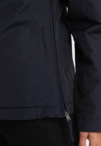 Veste bleu marine en tissu léger avec une fermeture éclair sur le côté. Elle possède une texture lisse et des détails cousus le long des coutures.