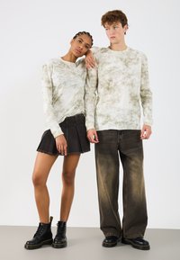 Długie rękawy koszulek w stylu tie-dye w kolorze zielonym i beżowym, noszone z czarną plisowaną spódnicą z denimu oraz szerokimi beżowymi dżinsami, połączone z czarnymi botkami.