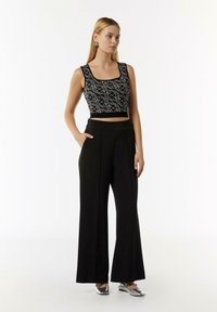 Schwarzes, kurzes Oberteil mit weißem abstraktem Muster, kombiniert mit hochtaillierten, schwarzen, weiten Hosen. Silberne flache Schuhe und minimale Accessoires.