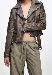 Bruine metalen cropped leren bikerjas met ritssluiting aan de voorkant en zijzakken, gecombineerd met licht olijfgroene broek met trekkoord.