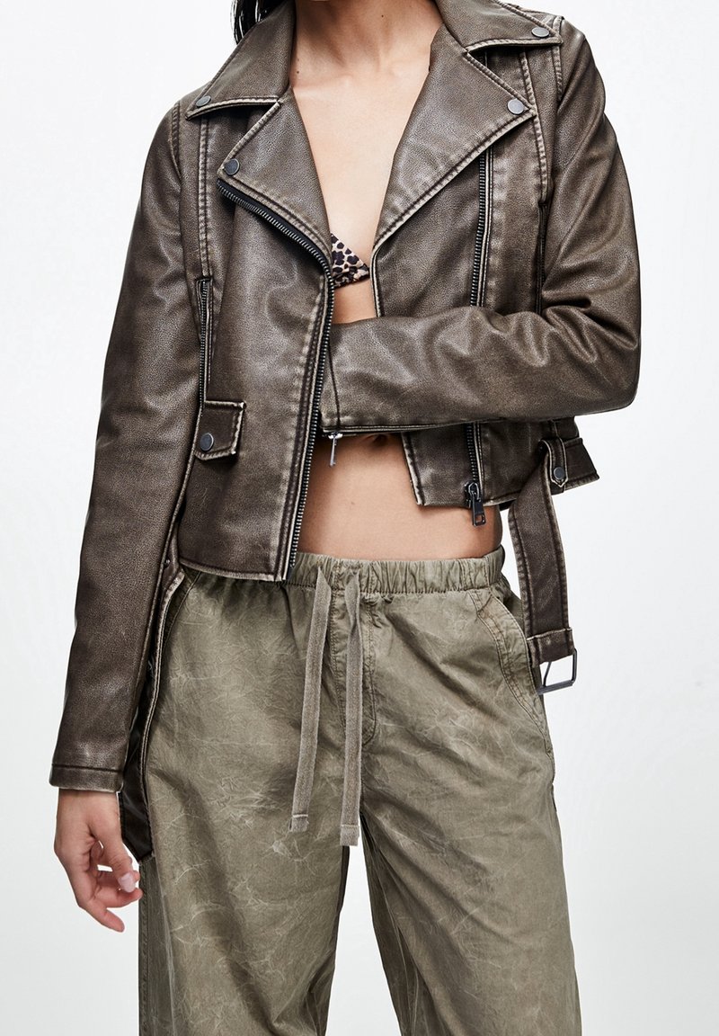 Bruine metalen cropped leren bikerjas met ritssluiting aan de voorkant en zijzakken, gecombineerd met licht olijfgroene broek met trekkoord.