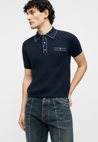 Άνδρας που φοράει κοντομάνικο πόλο σε ναυτικό μπλε με υφή και λευκή ραφή, καθώς και σκούρα μπλε τζιν, κοιτάζει αριστερά με το ένα χέρι στην τσέπη.