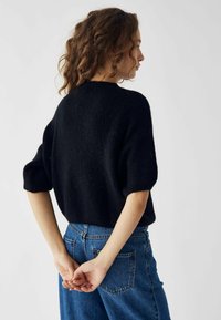 Čierny pletený sveter s krátkymi rukávmi, textúrovaná tkanina a uvoľnený strih, kombinovaný s modrými džínsami s vysokým pásom. Jednoduchý, ležérny dizajn.