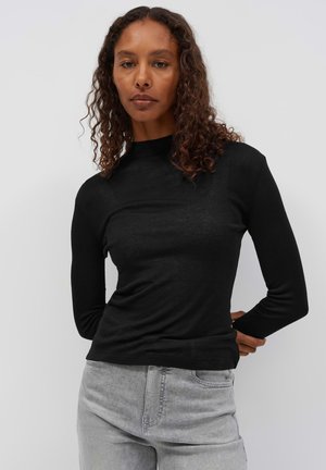 Donna con capelli ricci che indossa una maglietta nera a maniche lunghe e jeans grigio chiaro, in piedi con le mani dietro la schiena su uno sfondo semplice.