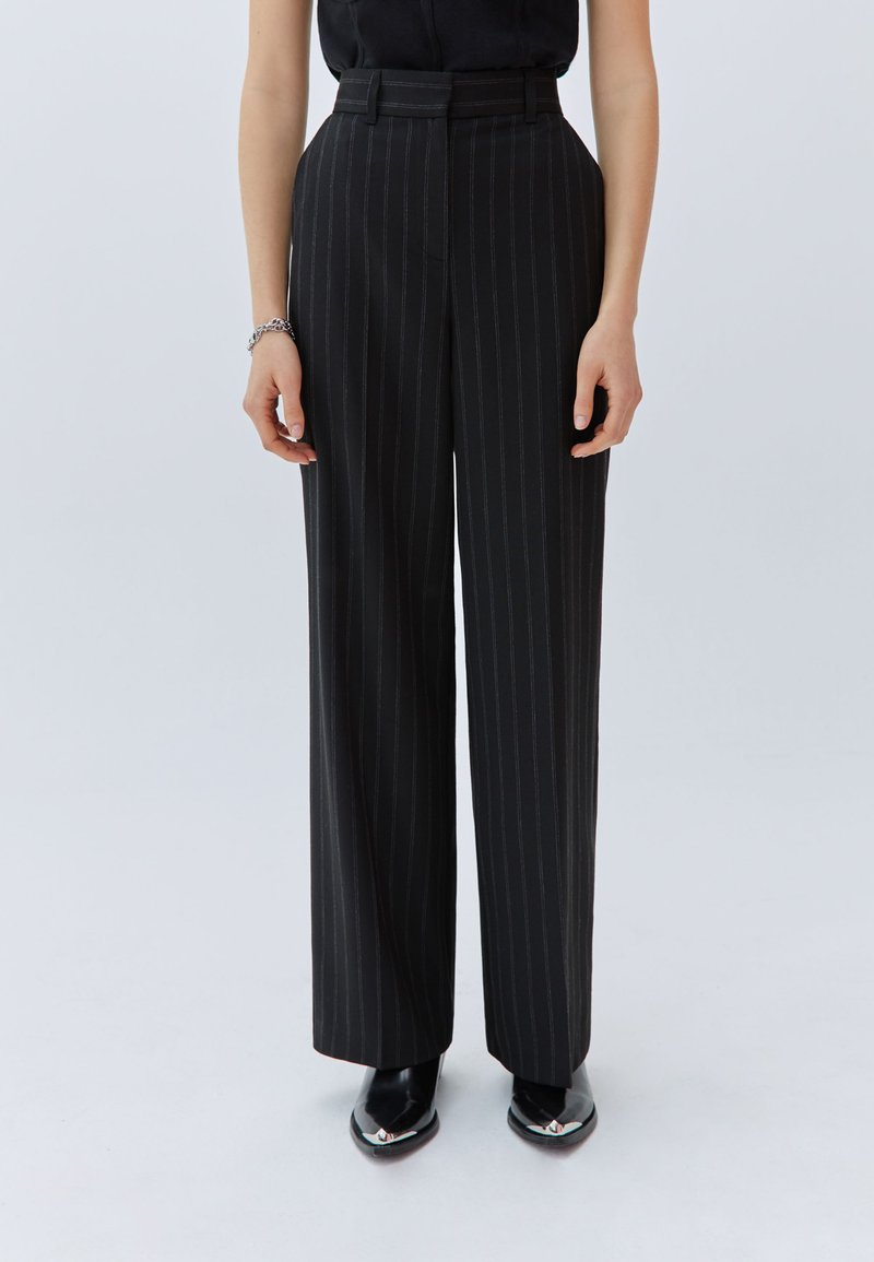 Pantalones de rayas negras y grises con cintura alta, pierna ancha y presillas para cinturón. Tejido suave y líneas limpias; combinados con zapatos negros de punta.