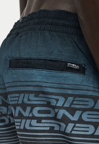 Κοντινή λήψη μπλε και μαύρων ριγωτών σορτς με ελαστική μέση και πίσω τσέπη με ετικέτα "O'Neill Hyperdry" σε άτομο με σκουρόχρωμο δέρμα.