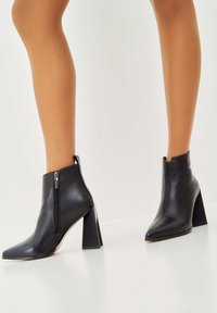 Černé kotníkové boty vyrobené z hladké kůže s špičatou špičkou a blokovým podpatkem. Zahrnují boční zip pro snadné obouvání.