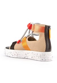 Sandales en matériau perforé beige, avec des accents orange et noir, une semelle blanche mouchetée et une fermeture éclair à l'arrière.