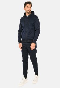 Felpa con zip di colore navy e pantaloni jogger abbinati, realizzati in tessuto morbido. Presenta un cappuccio, polsini a coste e tasche laterali. Indossato con sneakers bianche.