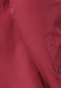 Roter Stoff mit glatter Textur, versehen mit einem Knopf und genähten Akzenten. Das Design zeigt eine einfarbige Oberfläche ohne Muster.