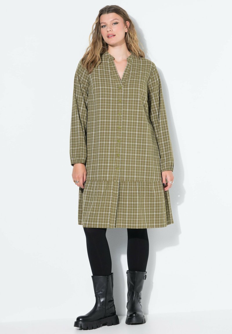 Olivengrünes kariertes Kleid mit durchgehender Knopfleiste, langen Ärmeln und schichtsaum. Kombiniert mit schwarzen Leggings und klobigen schwarzen Stiefeln.