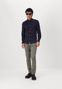 Jūrinis mygtukų marškinėliai slim fit stiliaus, pilki chinos ir juodi batai. Marškiniai turi apykaklę ir baltus mygtukus; modelis dėvi saulės akiniai.