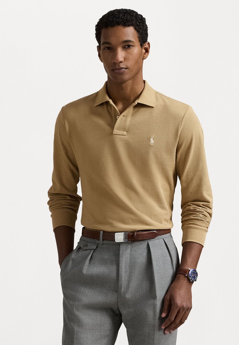 Ilgām piedurknēm polo krekls brūnā krāsā ar apkakles kakla izgriezumu. Valkāts ar pelēkiem plisētiem biksēm un brūnu jostu. Uz krūtīm ir logo.