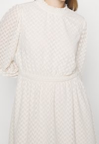 Vestido de crema texturizado con un escote alto y con volantes, y mangas largas. Presenta una cintura fruncida y un sutil patrón geométrico en toda la tela.