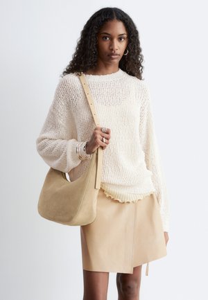 Kvinde med krøllet hår bærer en cremefarvet strikket sweater, beige lædernederdel, bærer en matchende beige skuldertaske og accessorizes med sølvsmykker.