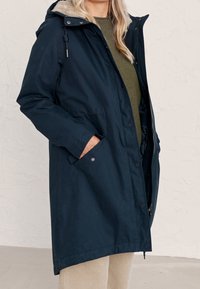 Granatowa parka z teksturowanej tkaniny, z kapturem wyłożonym futrem, przednimi kieszeniami i zapięciem na zatrzaski. Lekka i sięgająca do kolan.