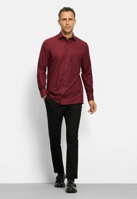 Bordeaux overhemd met lange mouwen en een zak, gecombineerd met zwarte broek en zwarte schoenen. Het overhemd heeft een gladde textuur.