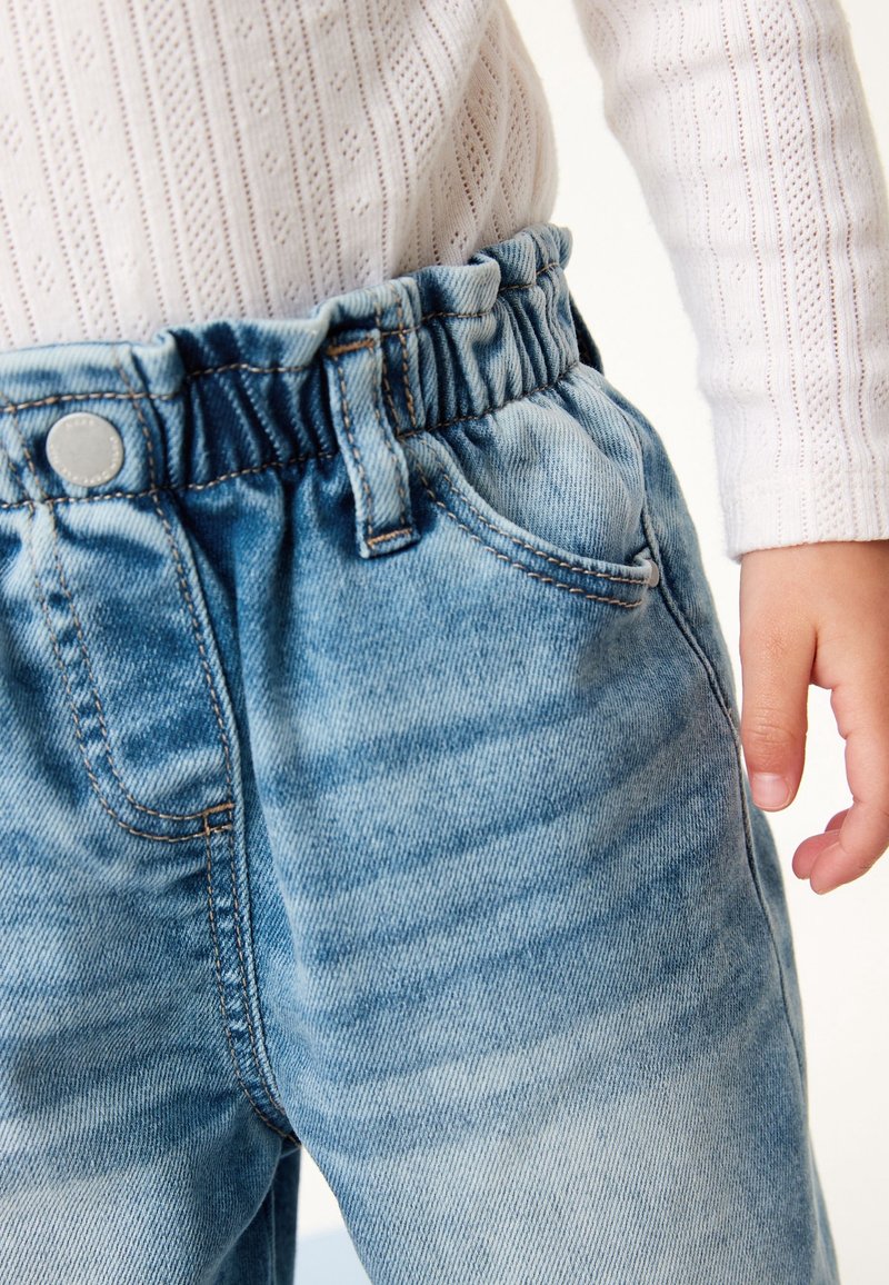 Enfant portant un pantalon en denim bleu clair avec une taille élastique et une chemise blanche texturée à manches longues.