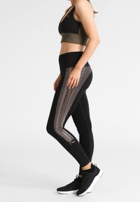 Leggings sportivi neri con un motivo texturizzato dorato lungo il lato, abbinati a un crop top nero con una fascia verde oliva. Scarpe da ginnastica nere.