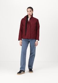 Burgundijas krāsas quilted jaka ar statīva apkakli, divām priekšējām kabatām un pogu aizdari, apvienota ar ziliem svītrainiem biksēm un melniem sporta apaviem.