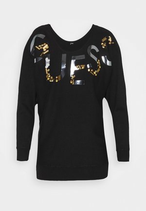 Pull noir à manches longues avec des lettres noires brillantes formant le mot "GUESS", ornées de perles dorées et noires sur la poitrine et les épaules.