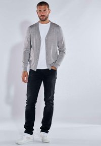 Grauer Cardigan mit Knopfverschluss, getragen über einem weißen Hemd, kombiniert mit schwarzen Jeans und weißen Sneakers. Glatte Textur und taillierte Passform.