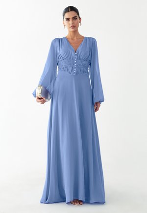 Frau trägt ein langes hellblaues Kleid mit Knöpfen und geraffter Taille, hält eine silberne Clutch und steht vor weißem Hintergrund.