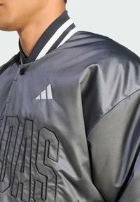 Gray satin bomber jacket with a black embroidered "ADIDAS" logo, featuring a ribbed collar with white stripes and snap-button closure.