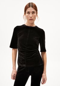 Zwarte fluwelen top met korte mouwen en een ronde halslijn. Heeft geplooide details aan de voorkant, een zachte textuur. Fittende pasvorm.