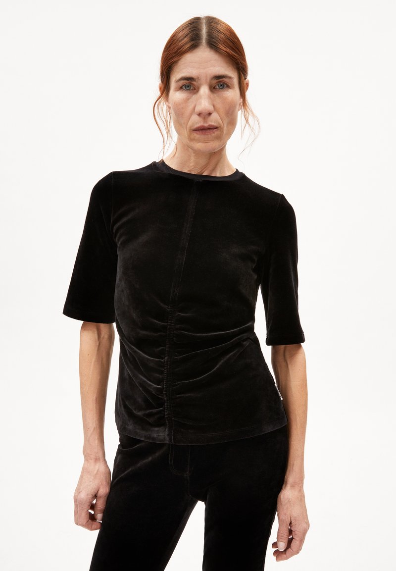 Zwarte fluwelen top met korte mouwen en een ronde halslijn. Heeft geplooide details aan de voorkant, een zachte textuur. Fittende pasvorm.