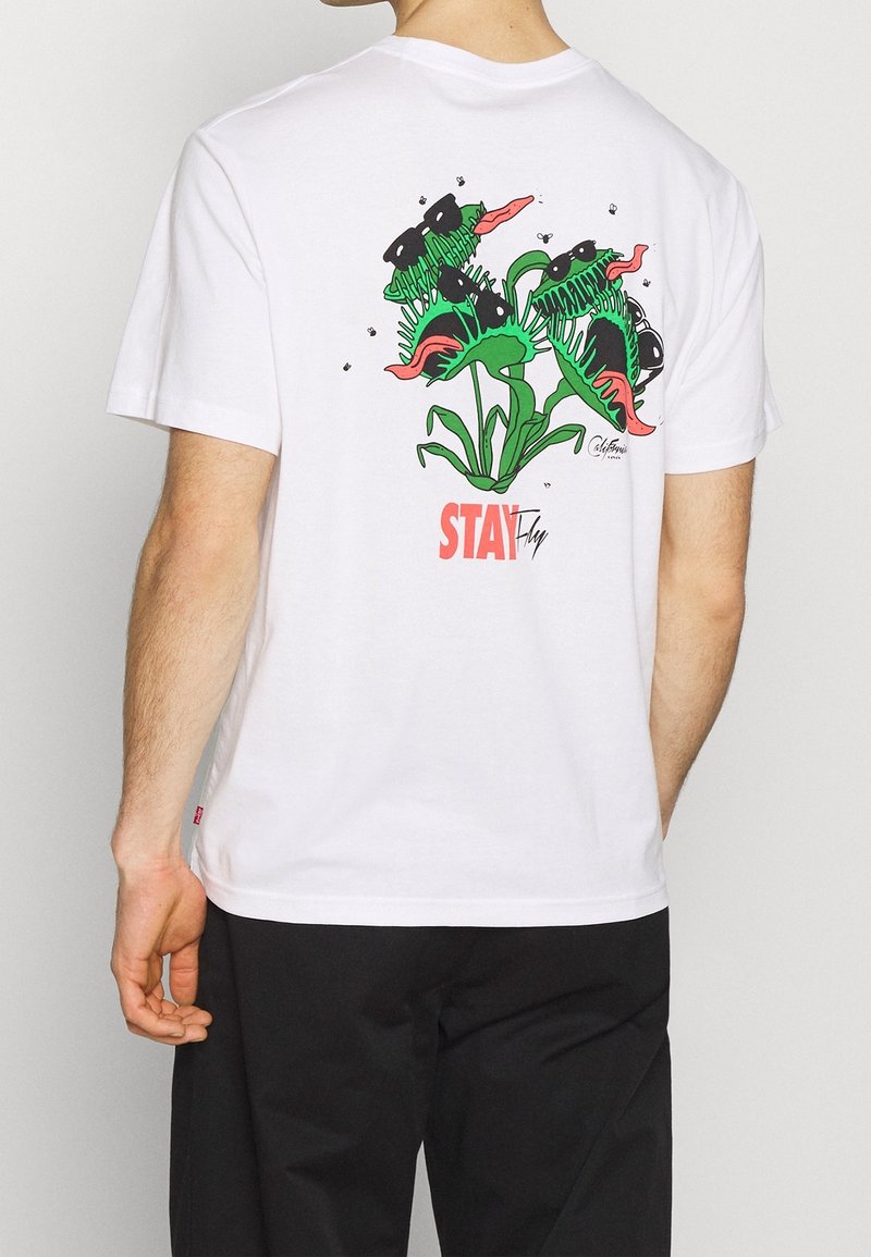 Valkoinen puuvillapaita, jossa on värikäs kasvi- grafiikka selässä, mustia, vihreitä ja vaaleanpunaisia yksityiskohtia, sekä teksti "STAY Fly."