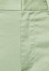 Pantalon vert clair en tissu texturé avec des lignes diagonales, comportant une poche arrière et des coutures piquées pour plus de détails.