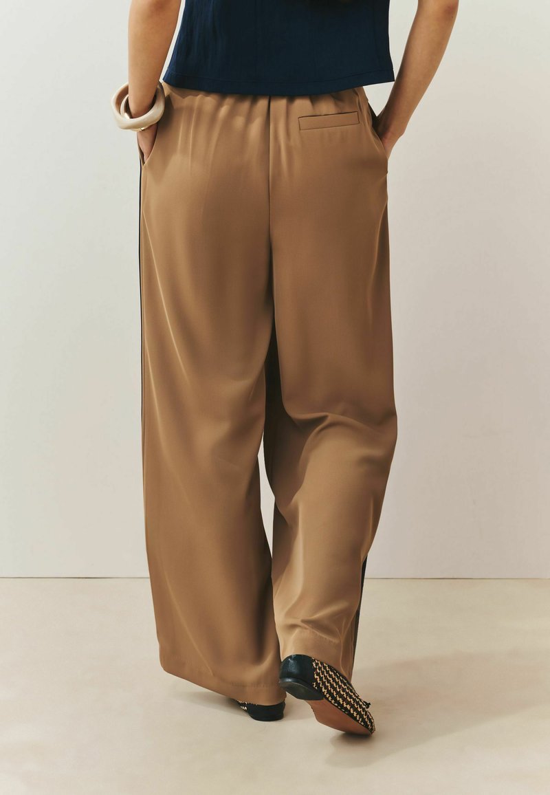 Personne portant un pantalon marron ample avec les mains dans les poches, des chaussures plates noires avec des semelles à motifs et un haut foncé.
