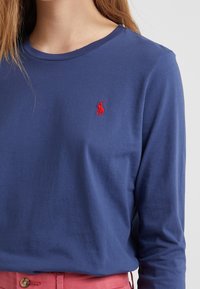 Modrá bavlněná trička s dlouhým rukávem, kulatým výstřihem a malým červeným vyšitým logem na hrudi; hladká textura, volný střih.