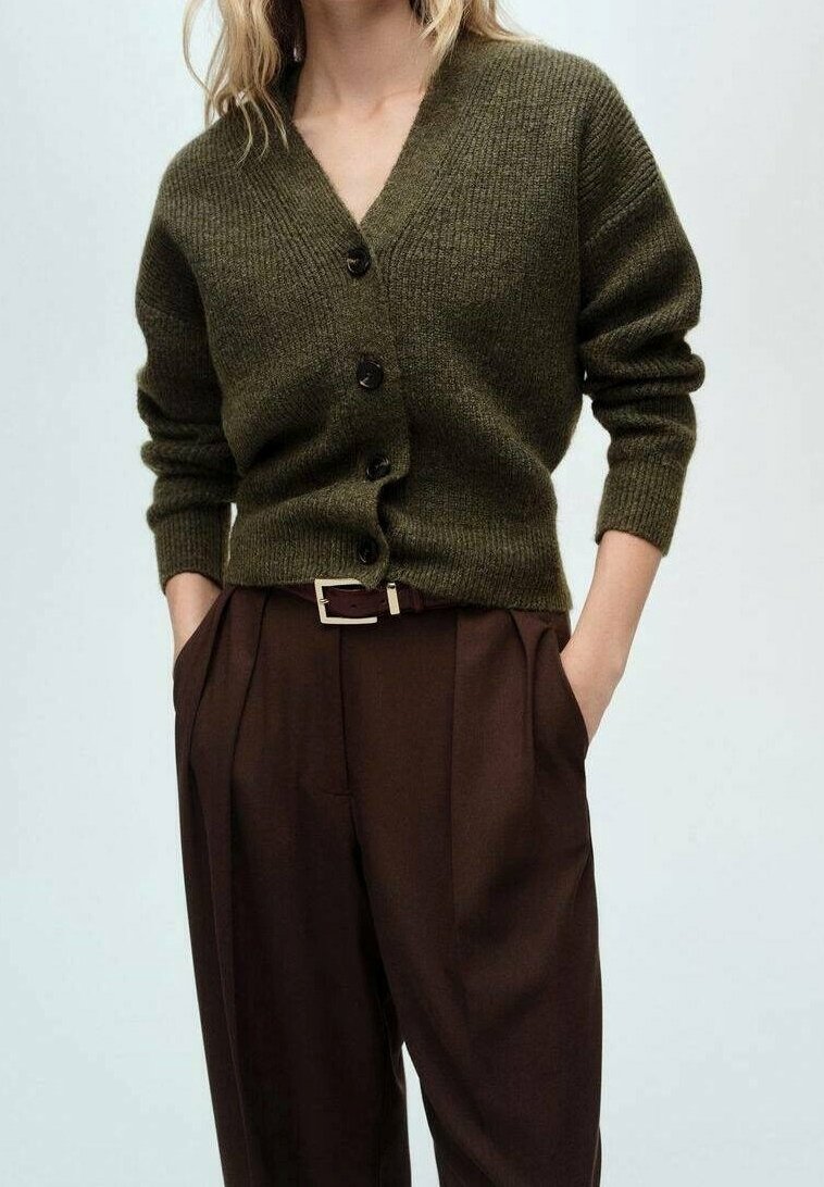 Femme portant un cardigan boutonné vert foncé rentré dans un pantalon plissé marron taille haute avec une ceinture, mains dans les poches.
