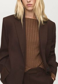 Brauner, taillierter Blazer mit einem strukturierten, gerippten braunen Pullover darunter. Der Blazer hat einen Reverskragen und einen einreihigen Knopfverschluss.