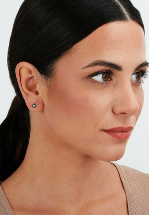 Runde Steckerohrringe mit einem türkisfarbenen Edelstein, eingefasst in einer Metallbasis, präsentiert an einem Modell mit dunklen Haaren und neutraler Kleidung.