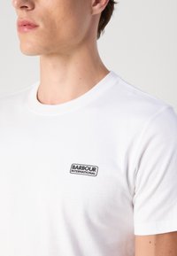 Fehér pamut póló, rövid ujjal és kerek nyakkivágással, a mellkasán fekete, téglalap alakú logóval, amelyen a "BARBOUR INTERNATIONAL" felirat olvasható.