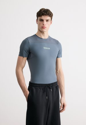 Mand iført tætsiddende grå kortærmet skjorte med teksten "Calvin Klein" og sorte bukser med snøre, stående med hænderne i lommerne.