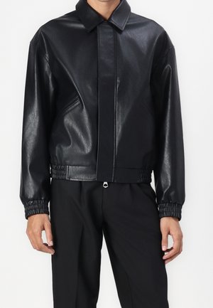 Chaqueta negra de cuero sintético con puños elásticos y cinturilla, usada con pantalones negros, cierre frontal con cremallera oculto bajo tapeta.