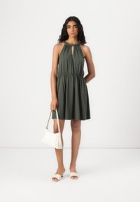 Vestido caqui sem mangas com decote em forma de buraco de chave e cinto elástico na cintura, combinado com uma bolsa branca em formato curvo e sandálias de slide bege.